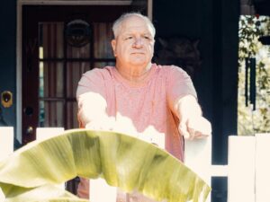 Senior man standing by a wooden fence, deep in thought, in a sunlit outdoor setting.