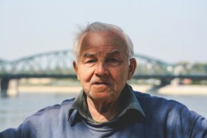 Portrait of an elderly man with gray hair in front of a scenic bridge on a bright day.