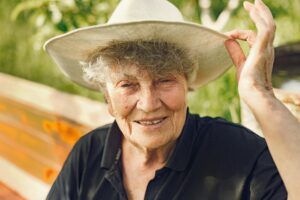 Portrait of an elderly woman wearing a sun hat, smiling under natural light in a lush garden setting.