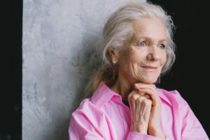 Close-up portrait of a smiling senior woman with blue eyes, hands together, wearing a pink shirt.