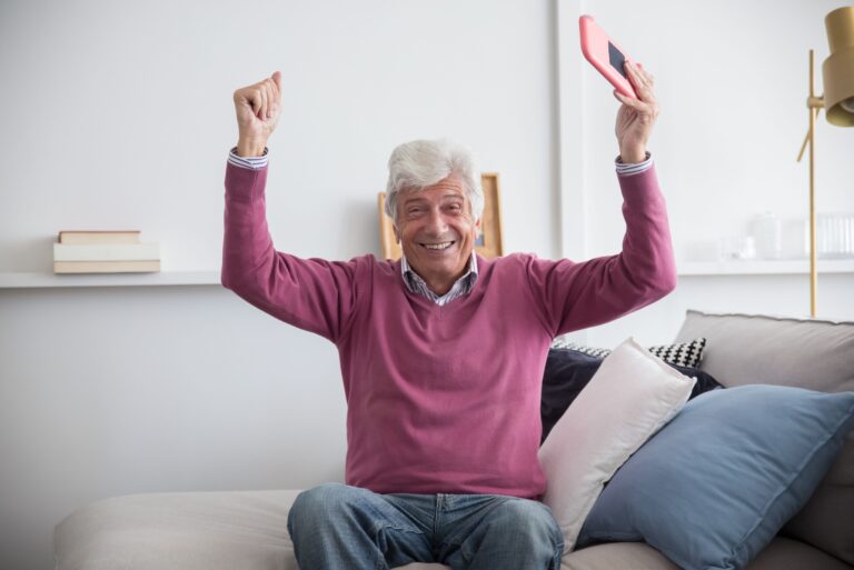 A happy senior man sitting on a sofa holding a smartphone indoors, celebrating with raised arms.