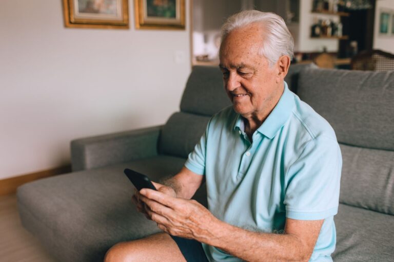 Elderly man in a polo shirt using a smartphone indoors while sitting on a sofa.