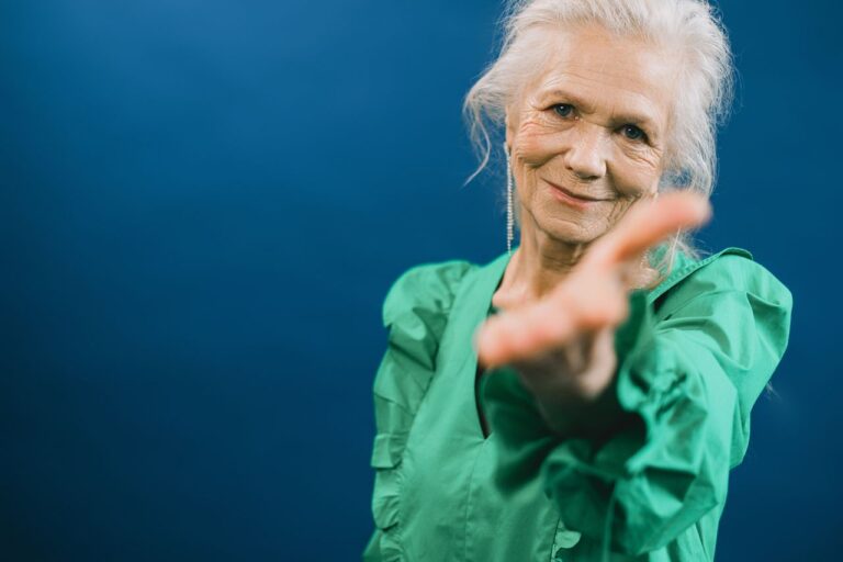 Elderly woman in green reaching forward with a smile against blue background. Studio portrait.