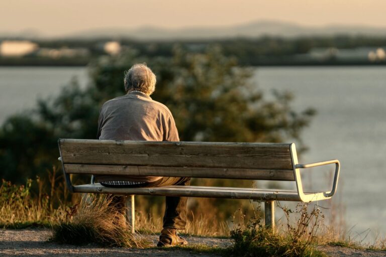 Serene scene of a senior man sitting on a bench by a lake during sunset.