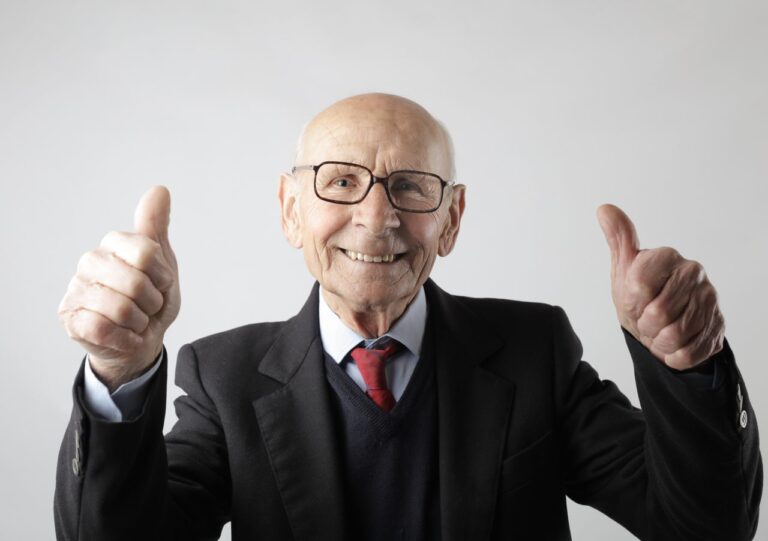 Optimistic senior man in elegant formal black suit and eyeglasses standing and showing thumbs up while smiling and looking at camera