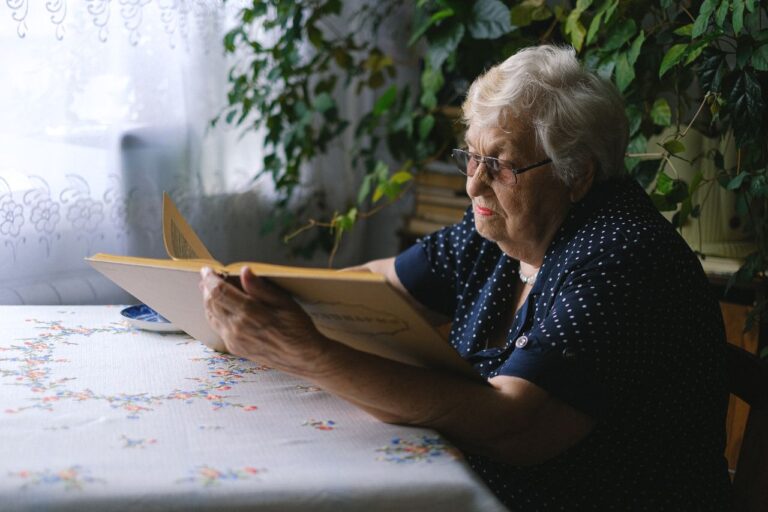 Side view of concentrated elderly female with gray hair and eyeglasses sitting at table and reading book at home with green plants