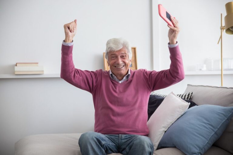 A happy senior man sitting on a sofa holding a smartphone indoors, celebrating with raised arms.
